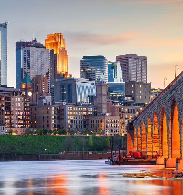 Downtown Minneapolis skyline at sunset with modern skyscrapers lit in golden light and the Stone Arch Bridge spanning the Mississippi River in the foreground.