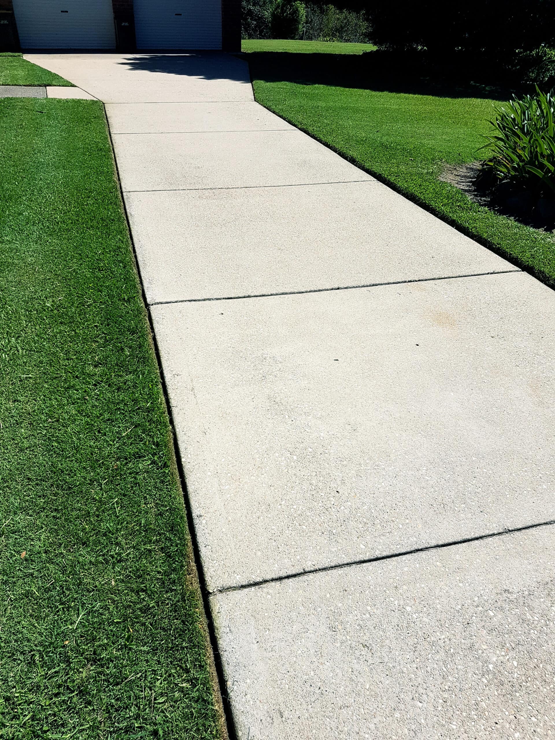 Concrete driveway with neatly trimmed green grass on both sides, leading to a double garage in the background on a sunny day.