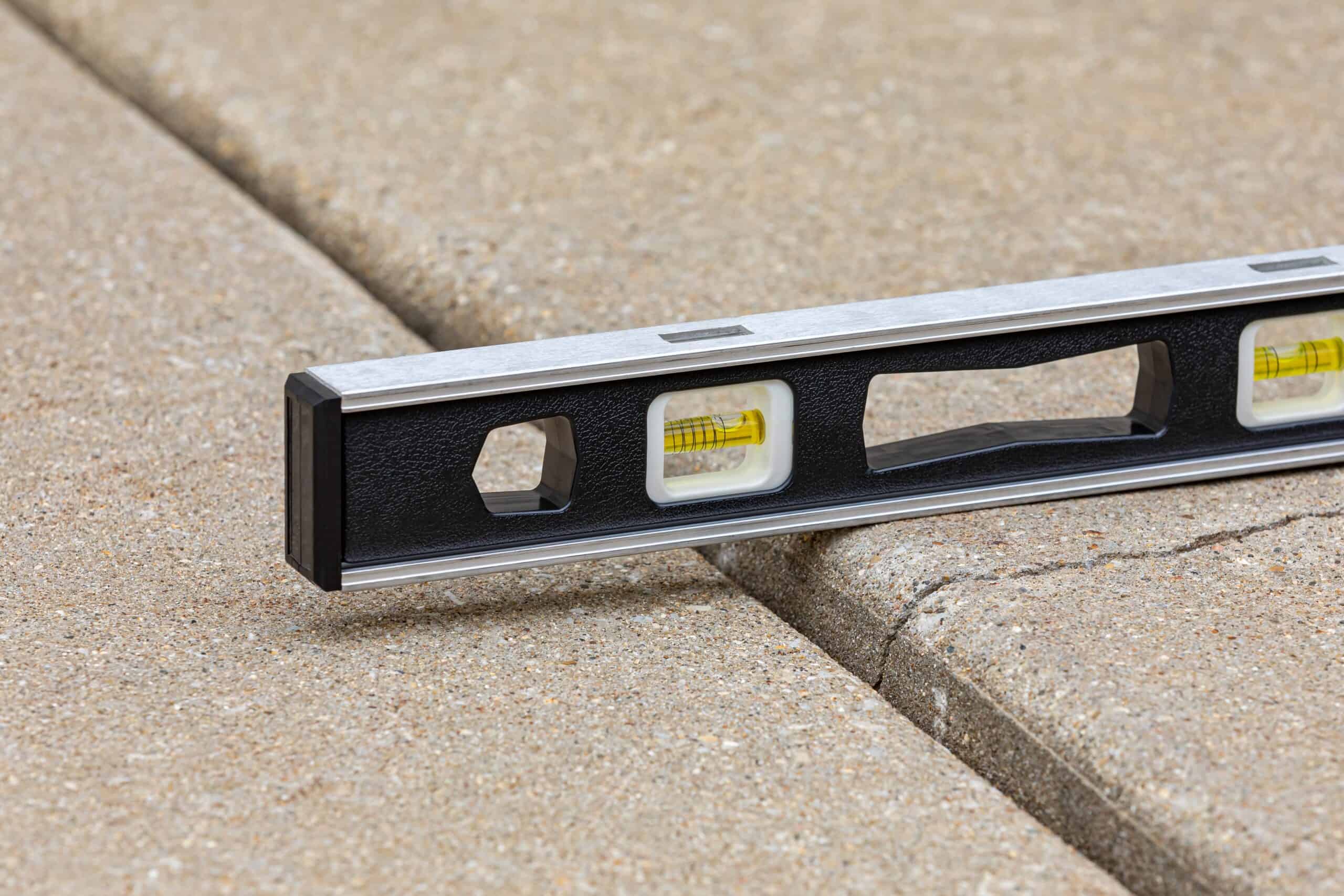 A spirit level placed across a raised concrete sidewalk shows uneven pavement, with one slab higher than the adjacent slab.