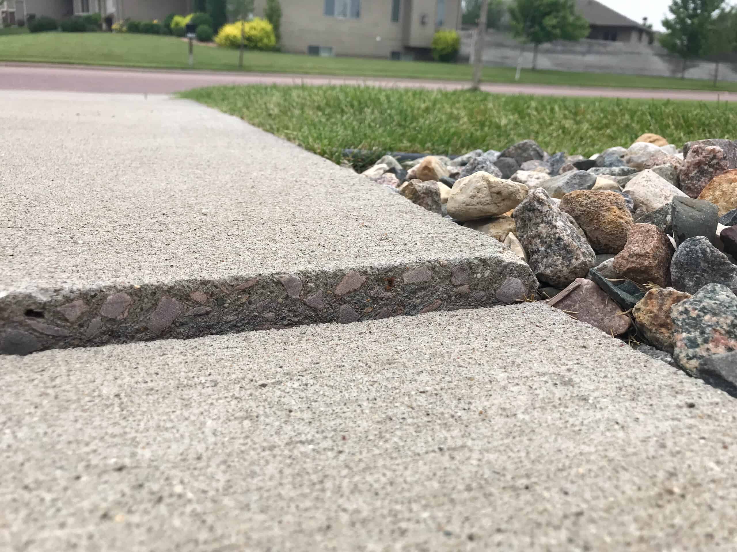 Close-up of a sidewalk with a raised concrete slab, creating a tripping hazard. Decorative rocks and grass are visible alongside the sidewalk, with houses and trees in the blurred background.
