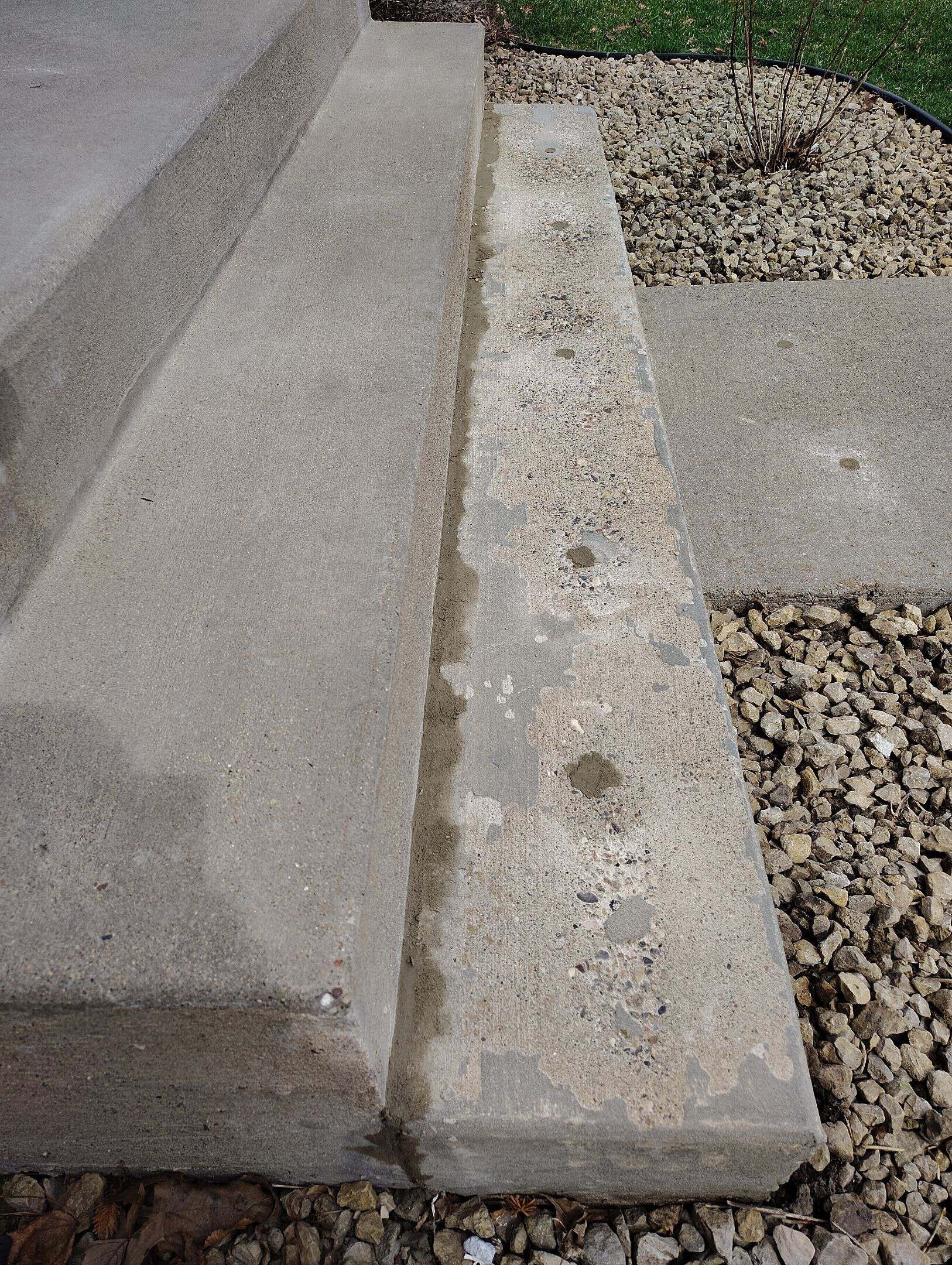 A concrete step next to a lower, weathered concrete slab with several small holes and patches; surrounded by rocks and a concrete walkway, with some grass and a bare plant in the background.