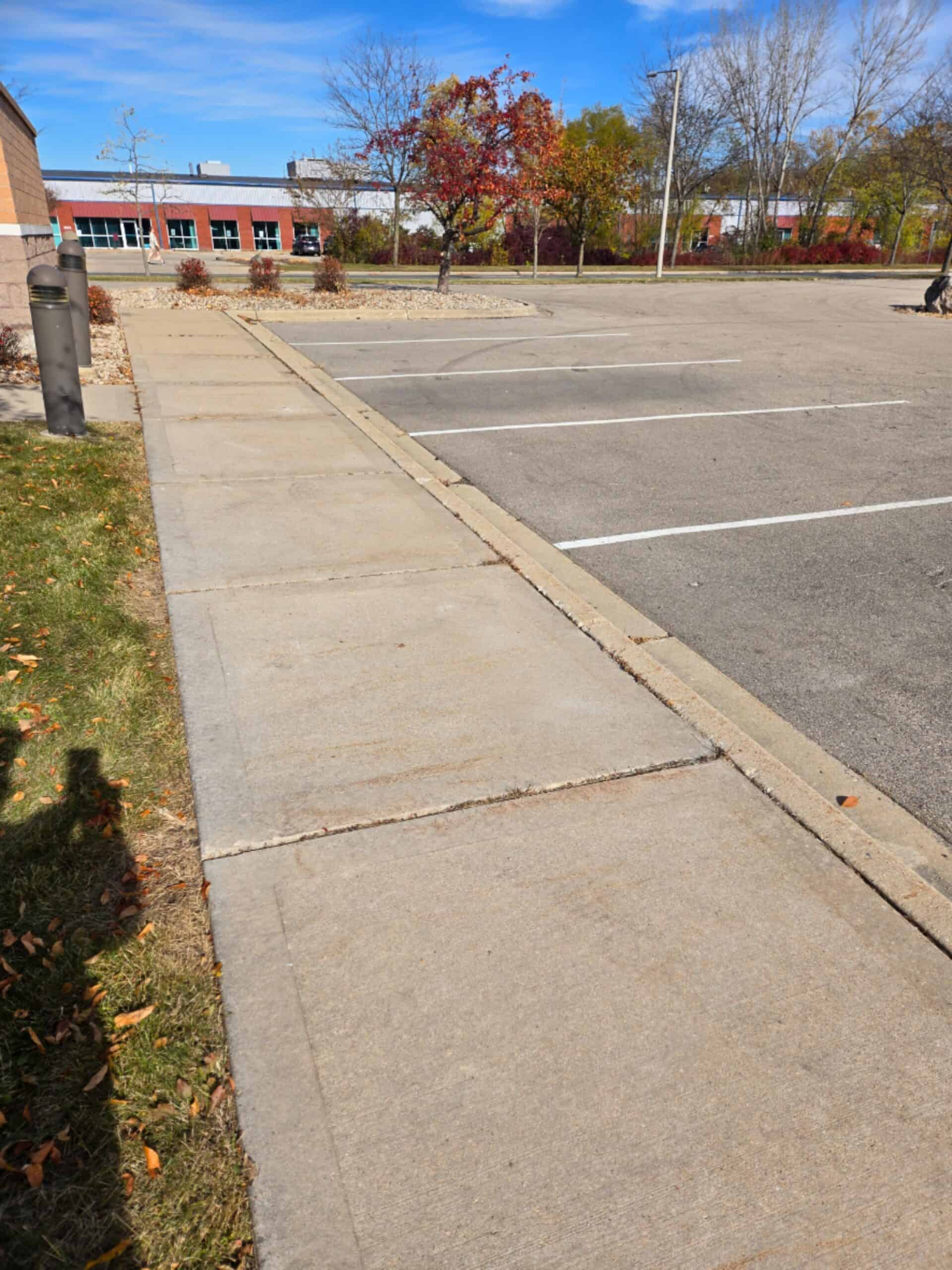 A sidewalk runs alongside an empty parking lot on a sunny day. Trees with autumn-colored leaves and a red brick building are visible in the background under a clear blue sky.