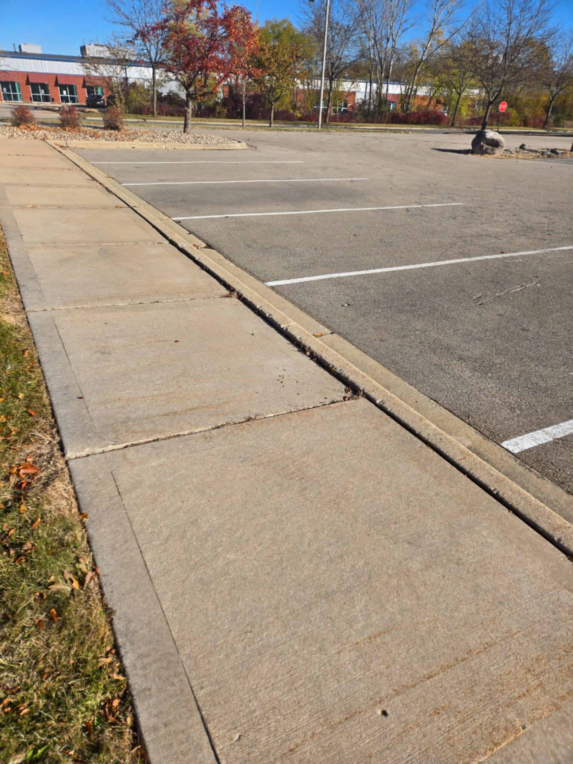 A concrete sidewalk runs alongside an empty parking lot on a sunny day, with a few autumn trees in the background and a red brick building visible in the distance.