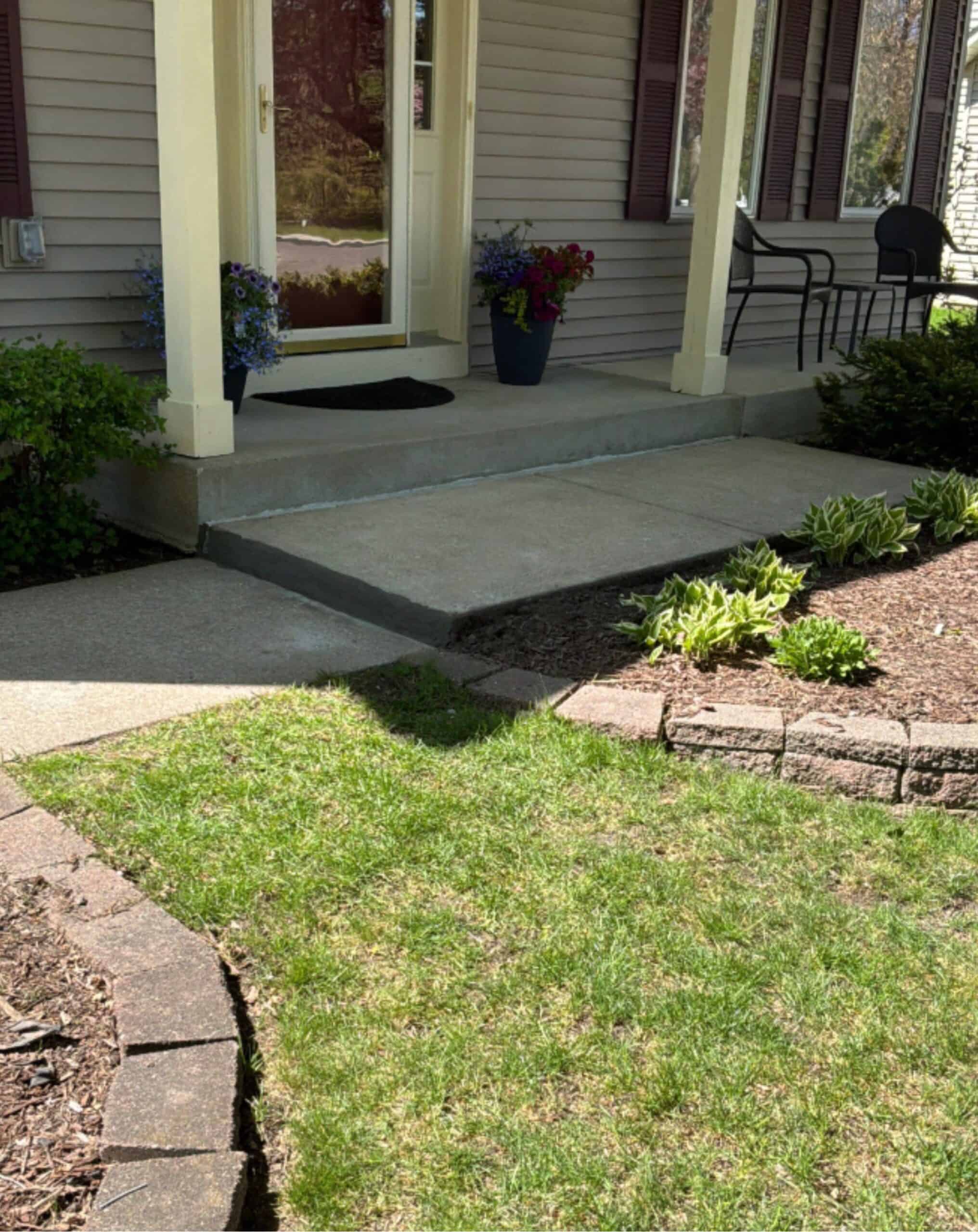 A concrete front porch in Minneapolis with two flower pots, a black doormat, beige pillars, and a black chair is bordered by green plants and mulch, with a curved brick edge and grass—a great candidate for mudjacking or polyjacking.
