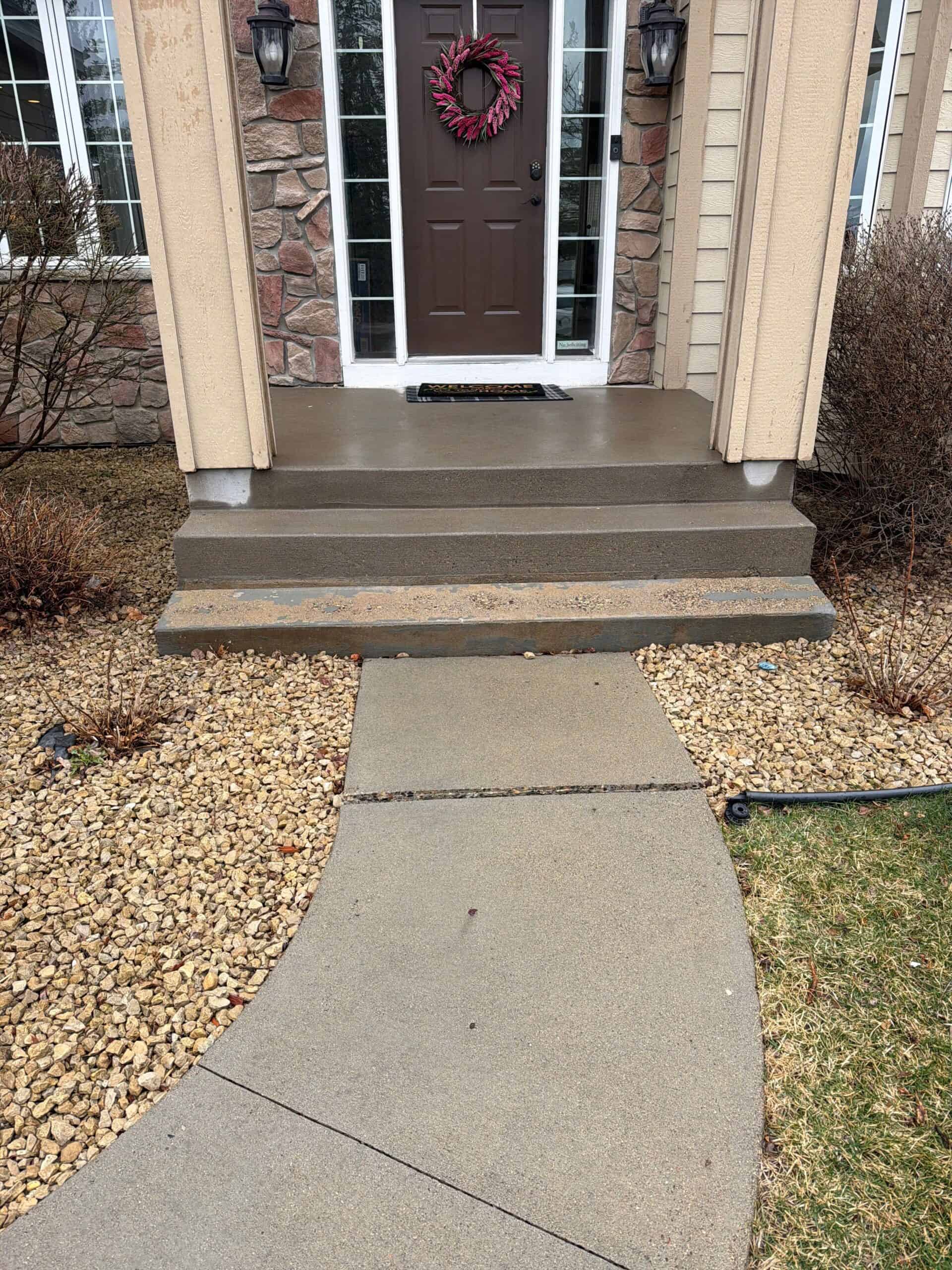 A concrete walkway curves toward a small front porch with three concrete steps, leading to a brown door with a wreath. Stone and beige siding, bushes, and rocks line the path.
