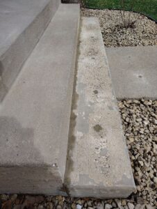 A concrete front step with surface damage and peeling, surrounded by rocks and a small concrete walkway. Some water spots are visible on the worn lower step.