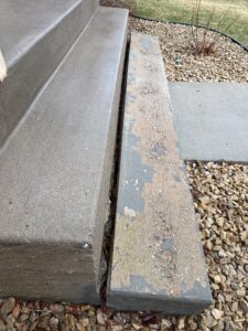 Worn, chipped concrete step beside smooth steps, bordered by rocks and a concrete walkway. Some grass and bare branches are visible in the background.