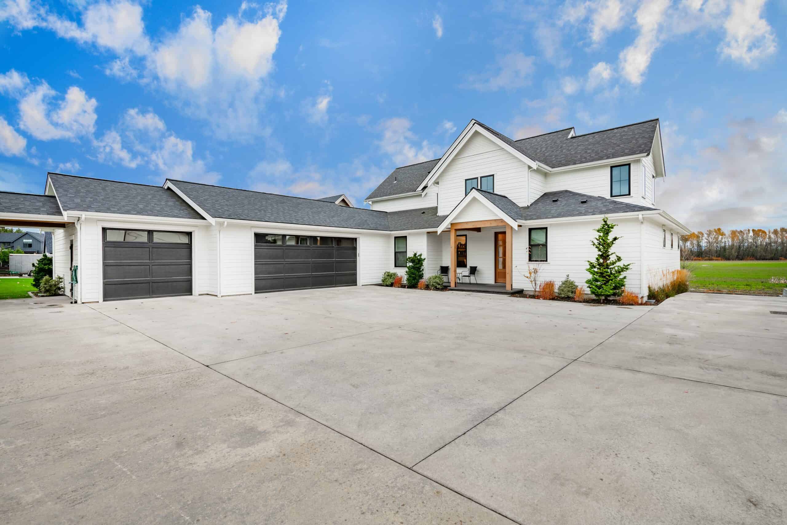Modern white two-story house with a large driveway, black garage doors, covered entryway, landscaped plants, and open green fields in the background under a blue sky with clouds.